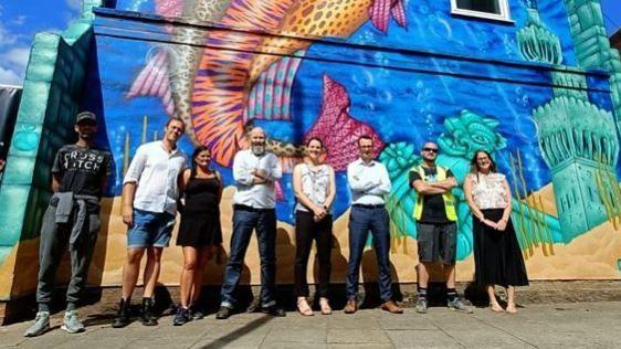A group of 8 people stand in front of a colourful mural on the side of a building. The mural depicts an orange and purple fish in an underwater setting with a turquoise coloured runed building in the foreground. The group is comprised of three women and five men.