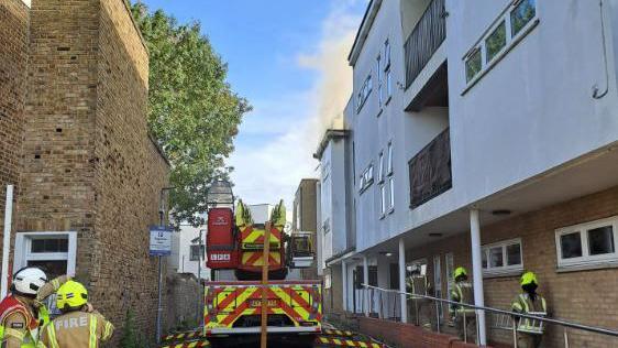 Firefighters in protective gear work at the scene of a blaze, with a fire engine parked between residential buildings and hoses laid out across the wet ground. Smoke is visible rising from the upper floor of a white building.