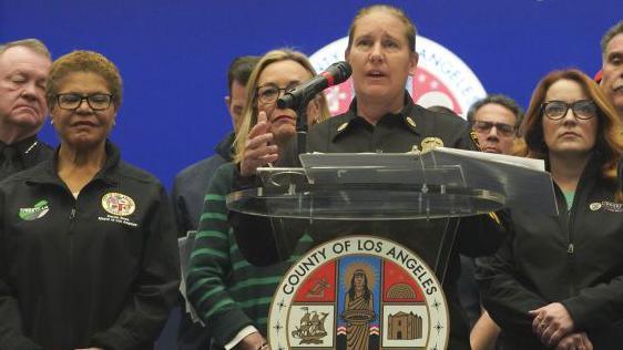 Crowley in black uniform with hair tied back at podium in front of people, including Karen Bass, with blue background