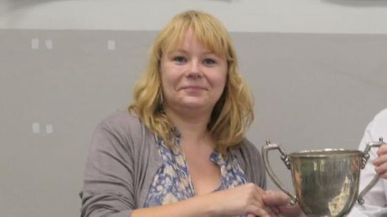 Kata Toma holding the Worcestershire Chess Championship trophy. She has blonde hair to her shoulders and a grey top over a blue and white dress.