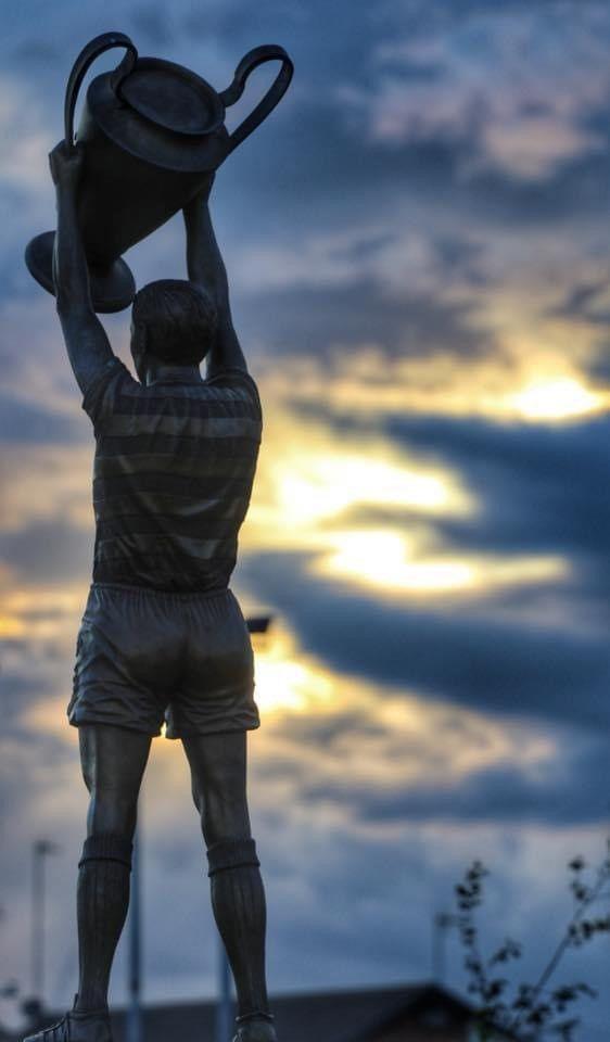 Statue of footballer holding a large trophy aloft, with a blue and yellow sunset sky in background.