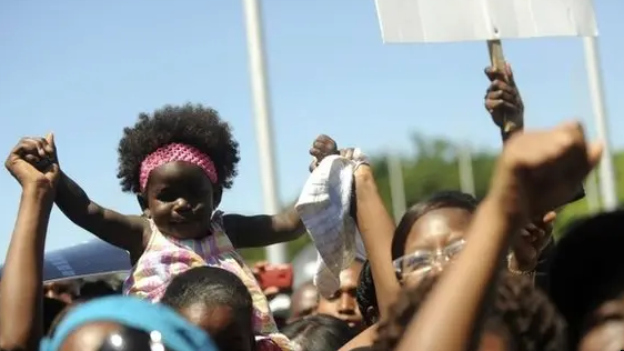 People protest against a constitutional court ruling in the Dominican Republic that redefined citizenship to exclude children of undocumented migrants, mostly of Haitian descent. In the foreground, a young black girl with her arms raised by one of the protesters sit prominently on someone's shoulders.