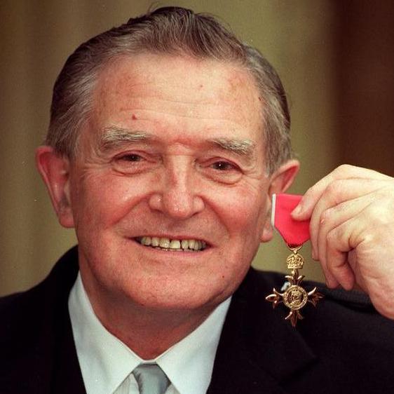 Jimmy Logan, in formal dress with white shirt and pale grey tie and black jacket, is holding a red banded OBE medal and smiling.