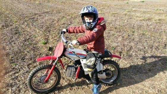 A young mason sitting on a scrambler-style bike in a field. He is wearing a read coat and a helmet, as well as black leather trousers. He is looking into the camera. 