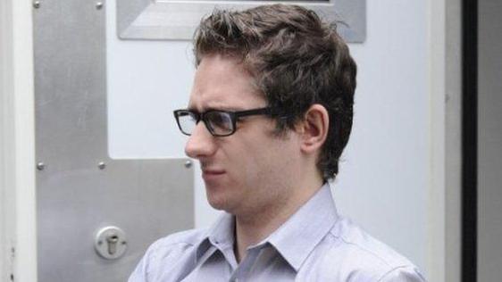 A man with dark hair and glasses, wearing a light-coloured shirt, in a close-up shot. He is walking past a heavy security door