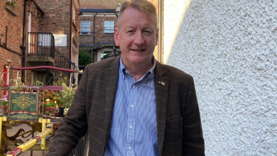 Jim Roddy is wearing a tweed blazer and blue and white striped shirt. 
He has short brown hair and is standing near a large white gable wall and a floral arrangement.