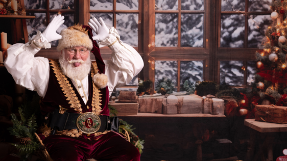 Allan Evans dressed as Santa, sticking his tongue out with his hands in the air, in front of a snowy window background and a Christmas tree