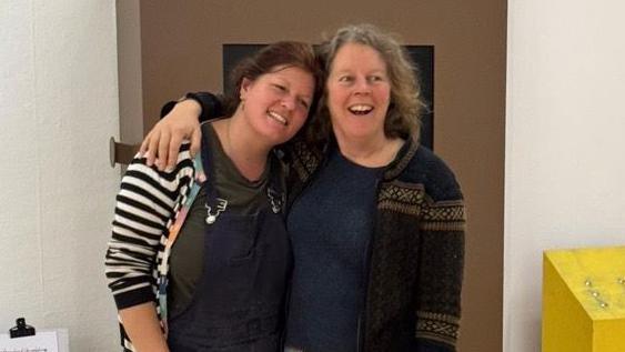 Jeni Cairns and Karen MacKelvie standing arm in arm in front of a brown wall inside. Jeni is wearing navy overalls over the top of a black and white striped T-shirt with long sleeves. She has brown hair tied behind her head and is smiling. Karen is wearing a brown knitted jumper over a navy top. She has shoulder length, light brown hair and has her mouth open in excitement.