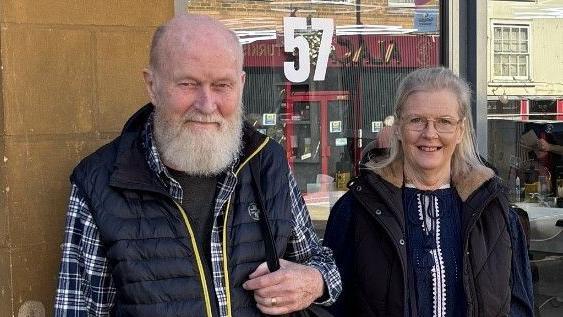 Peter on the right of this image is shown after a recent hair cut back in the UK. His beard is trimmed and his hair shorter. His wife, Barbie, stands smiling next to him.