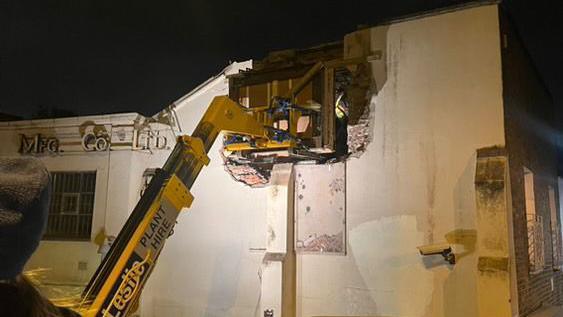 At night, a boom lift is used to remove part of an the building’s wall, revealing its interior. A worker in a high-vis jacket is inside. 