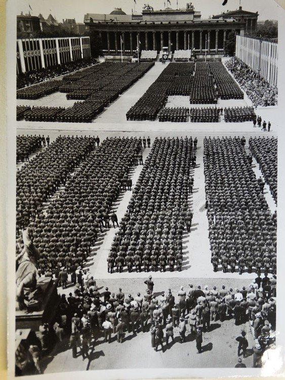 Large numbers of troops fill through a broad plaza in strict formation in Berlin at the Condor Legion homecoming. In the distance, Nazi banners can be seen