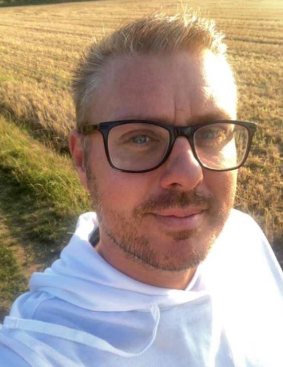 The photo shows a man smiling at the camera. He's wearing black frame glasses, and has blonde spikey hair. He's also wearing a white hoodie. He's standing in front of a field. 