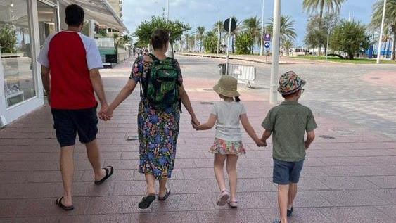 A man in a red and white t-shirt holds hands with a woman wearing a flowery dress, a girl and a boy as they walk down a street with palm trees in the background. All of them have their backs to the camera.