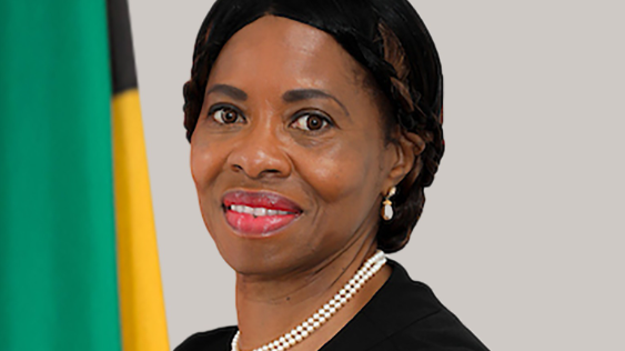 Dr Beverly Lindsay looks at the camera and is wearing a beaded necklace and earrings. The flag of Jamaica is in the background.