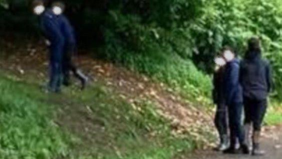 Five young children at a grassy slope in a park near trees. Three stand on a path and two are on the slope. All have their faces blurred.