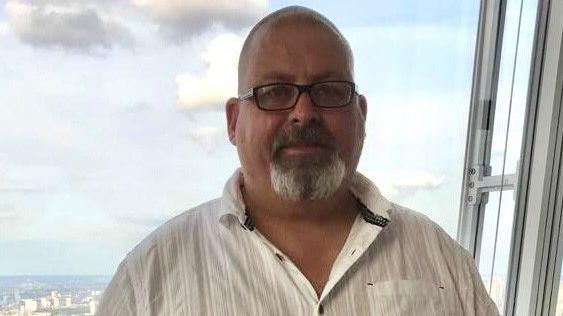 Mark Randle looking at the camera. He is in front of a window showing a view of a town or city and he wears a white shirt and has a short goatee beard and wears glasses with a shaved head