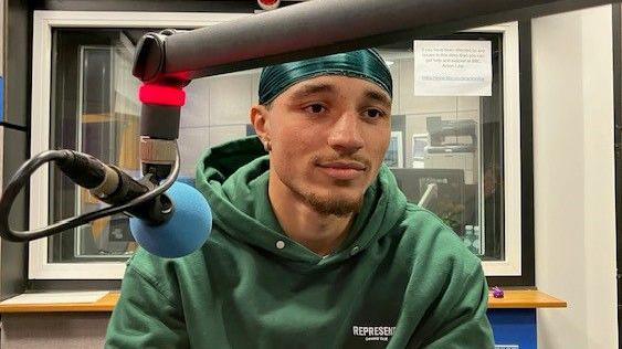 Issiah Hamilton-Allen sitting in a radio studio with a microphone infront of him, wearing a green hoodie and a green bandana