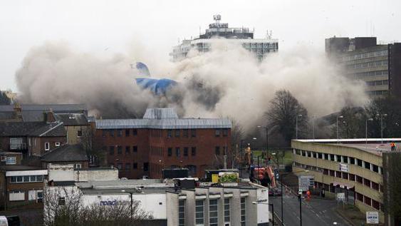 Clouds of smoke arising from the demolition of a building which is hardly visible apart from a section covered by a blue sheet. There is a smaller brick building in front and a concrete car park to the right.