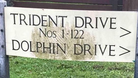A white rectangular sign fixed to two grey posts. The words "TRIDENT DRIVE Nos 1-122" and  "DOLPHIN DRIVE" are written on the sign.

