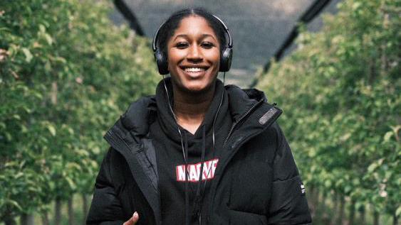 Thaliqua Smith stands smiling at the camera, with hedges either side of her. She has her black hair tied up and is wearing a pair of black headphones. She has a black Marvel hoodie on, with a black puffer jacket over the top.