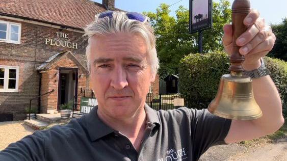 Adam Nicoll, ringing a pub bell, while standing outside The Plough, King's Walden 