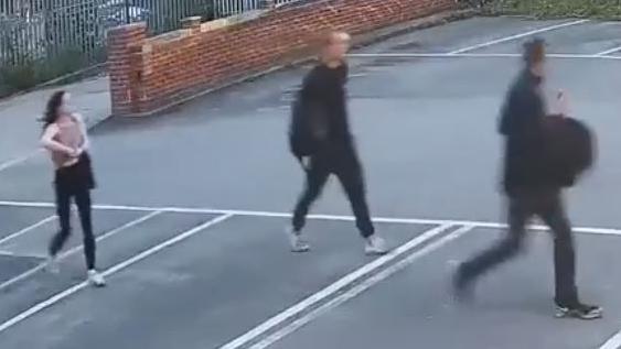 A blurry CCTV still of three people walking across what appears to be a car park. Two of the people are wearing dark clothing while the third, appearing to be a woman, is wearing a pink top and appears to be holding an item in her arms.