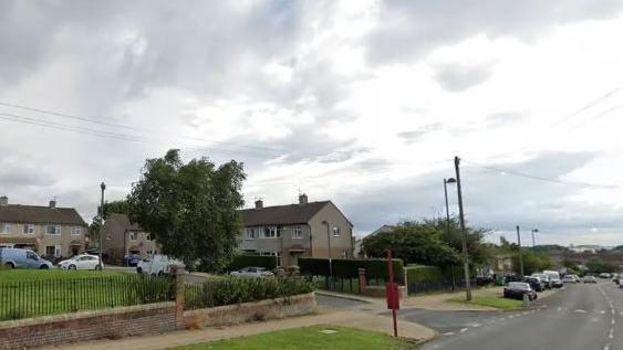 An estate in Holme Wood, Bradford. The main road is empty, with a side road containing several parked cars. A red bus stop post is in the foreground. 
