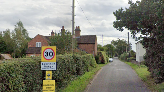 A country lane with grass on either side of the road and a red brick property to left and a cream smaller house on the right. There is a road sign before the house that reads "30mph" and an area sign that reads "Edgmond Marsh". 