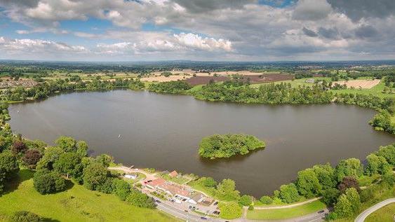 A wide aerial picture of a large lake with a small tree-filled island in it. The lake is surrounded by trees, bushes and green fields as well as farmland.