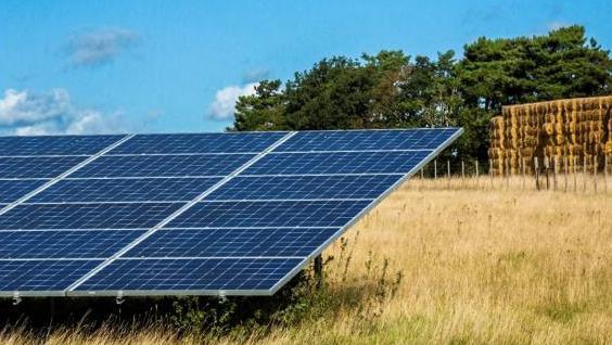 A row of solar panels in a field with a stack of bales and trees in the background.