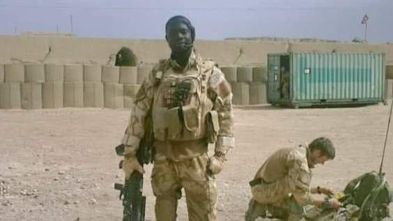 OB poses for a photograph at a camp in Afghanistan. He is wearing camouflage kit and carrying a large gun by his side.  