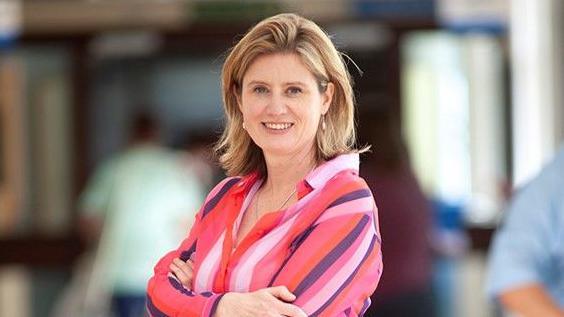 Dr Frankie Swords is standing to the side looking towards the camera. She is wearing a multicoloured striped shirt, and has arms crossed over her chest. She has shoulder length blonde hair and is smiling at the camera.
