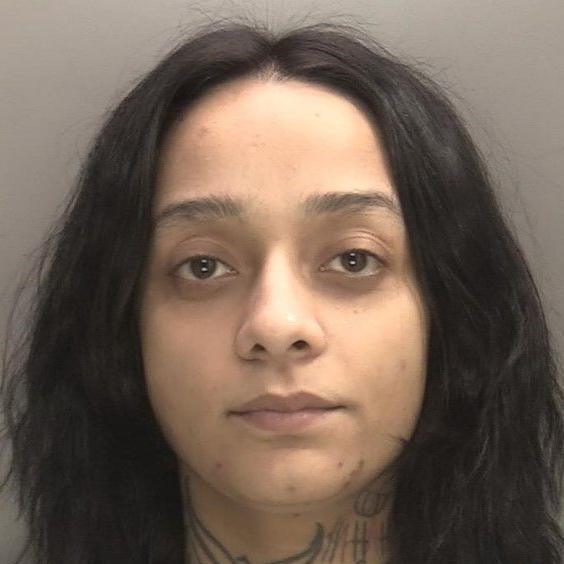 A mugshot of Jaskirat Kaur, who is staring straight at the camera. She has brown eyes and dark hair, and tattoos on her neck. The photo of her is a close-up of her face and neck and she is in front of a grey background.