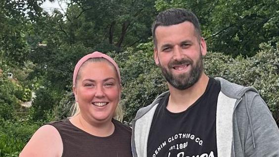Sophie Cole, who has her blonde hair tied back by a pink headband. She is wearing a black vest and is smiling. She is standing next to her partner, who has short black hair, a beard and is wearing a black T-shirt and grey jumper. They are standing in front of trees.