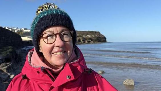 A woman is wearing a red winter coat, with a two-tone dark blue and marine woollen hat with a yellow and black and white pom-pom. She is wearing glasses and smiling. Behind her is part of Benllech beach and sea, and a part of the Anglesey coast headland on the horizon.