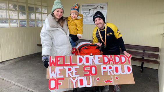 Jered and his wife hold a cardboard sign saying 'Well done daddy, we're so proud of you' with hearts and a moustache on. His wife is holding their young baby. They're wrapped up for the cold weather.