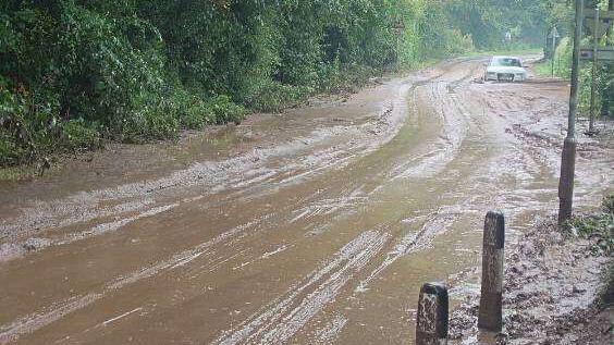 A really muddy road, with a car in the distance and one side lined by a tall hedge, and bollards on the other