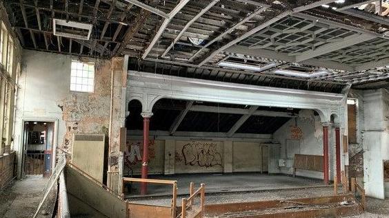 The inside of the building with stripped peeling walls, ceiling rafters and a stage.