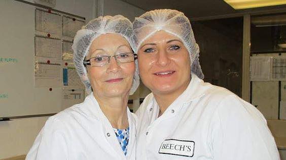 Barbara Collins (left) and Monika Nejman (right) wearing white Beech's overalls and white hair nets with their heads touching in Beech's factory. They are both smiling.