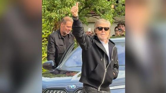 Paul McCartney waves to the crowd. He is smiling and wearing sunglasses. He is wearing a light coloured top, black jeans and a black hooded jacket. He is standing in front of a car and some people can be seen in the background, including someone filming on their mobile phone.