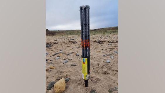 A large firework canister. The black plastic tubing is long and tall. Its pointed base is stuck in the sand. It looks like it can hold several fireworks. Rocks are also strewn among the sand.