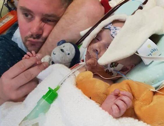Chris Cookson lies with his arm resting on his head and his finger holding the tiny hand of his son Carter. Carter is lying next to him with a tube coming form his mouth and a blanket draped around his body and head.