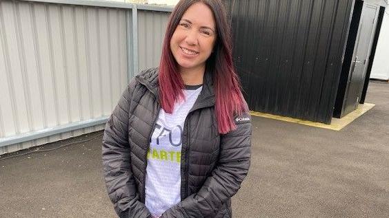 Michelle Hume with brown hair dyed red at the ends and wearing a white top with a logo and a black padded coat, stands in front of  black metal shed and cream metal fence.