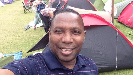 A man in a navy polo shirt taking a selfie. He is standing in front of a line of tents.