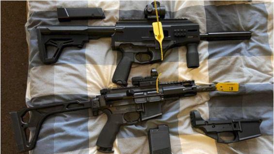 Two black assault rifles with a number of accessories placed on a bed with a blue and white duvet