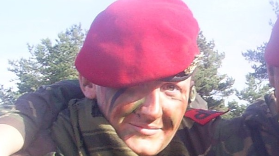 A woman in a red beret, with smears of camouflage paint on her face, and wearing a camouflage top, looks at the camera