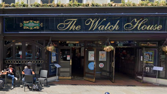 Picture of The Watch House pub with signage written in gold on a blue frontage. Outside the pub there are hanging baskets and a few people sat the left with drinks. 