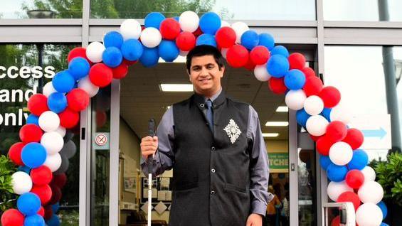 A man wearing a grey shirt and black waistcoat stands in front of a red, white and blue balloon arch and holds a white cane.  Behind the arch is a doorway which is open, showing the inside of a building behind.