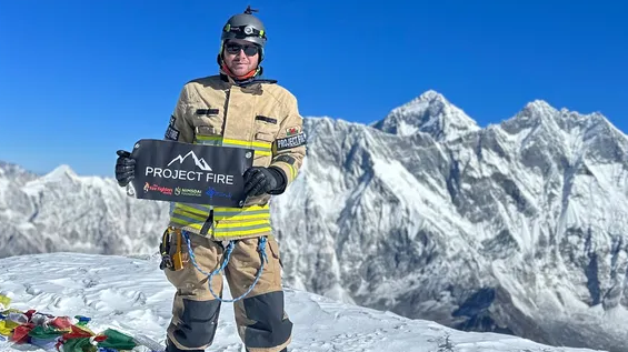 Rhys Fitzgerald in his firefighter gear standing on a mountain, with mountains behind him. He's holding a sign saying 'Project Fire'