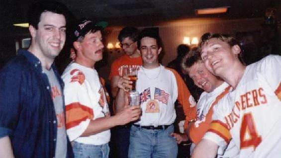 Gareth and a group of other Bucs fan at a meeting. All are looking at the camera and smiling. They are wearing Bucs merchandise, which was orange and white. 